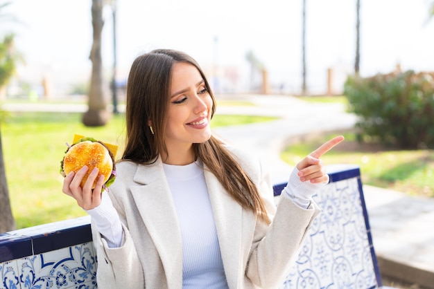 Young pretty woman holding a burger at outdoors pointing to the side to present a product