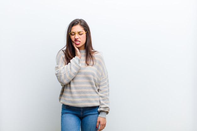 Young pretty woman holding cheek and suffering painful toothache, feeling ill, miserable and unhappy, looking for a dentist isolated against white wall