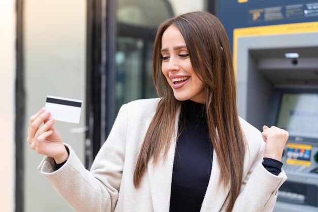 Young pretty woman holding a credit card at outdoors celebrating a victory