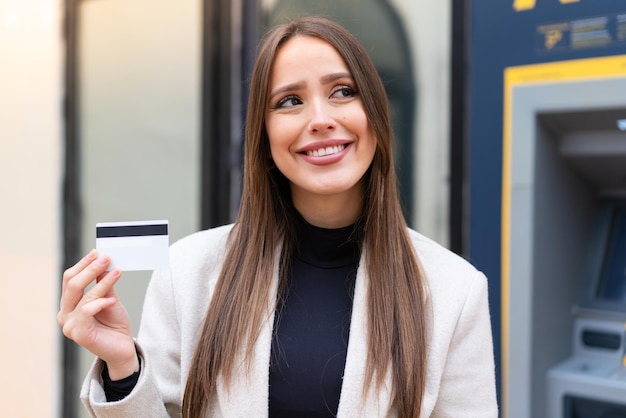 Young pretty woman holding a credit card at outdoors looking up while smiling