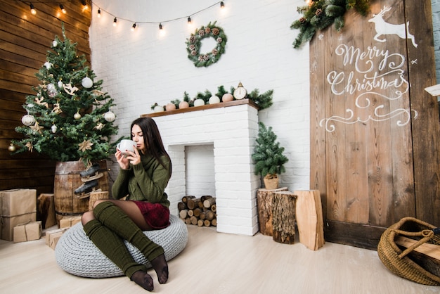 Young pretty woman holding a cup with a hot drink with Christmas tree on background