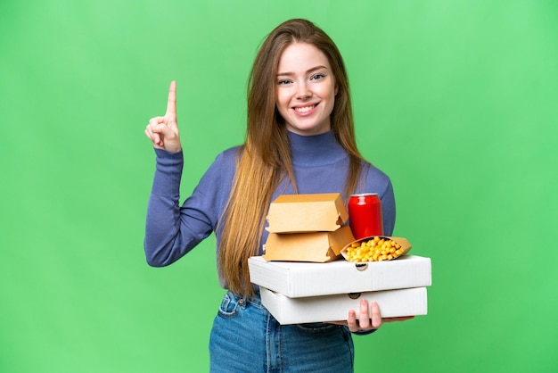 Young pretty woman holding pizzas and burgers over isolated chroma key background pointing up a great idea