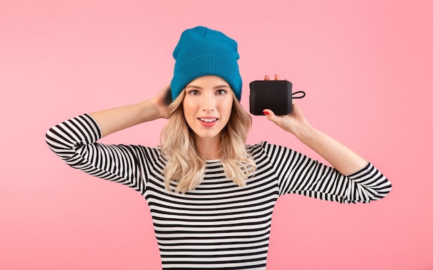 Young pretty woman holding wireless speaker listening to music wearing striped shirt and blue hat smiling happy positive mood posing on pink background isolated