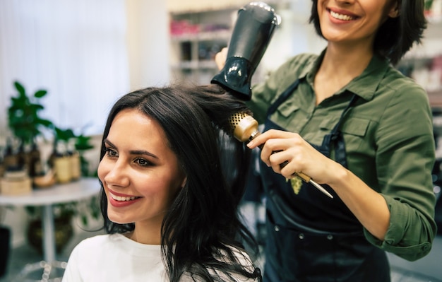 Young pretty woman is sitting in the armchair in a beauty salon getting her hair dried by a smiling professional stylist.