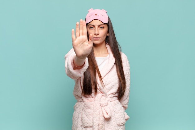 Young pretty woman looking serious, stern, displeased and angry showing open palm making stop gesture. awaking wearing pajamas concept