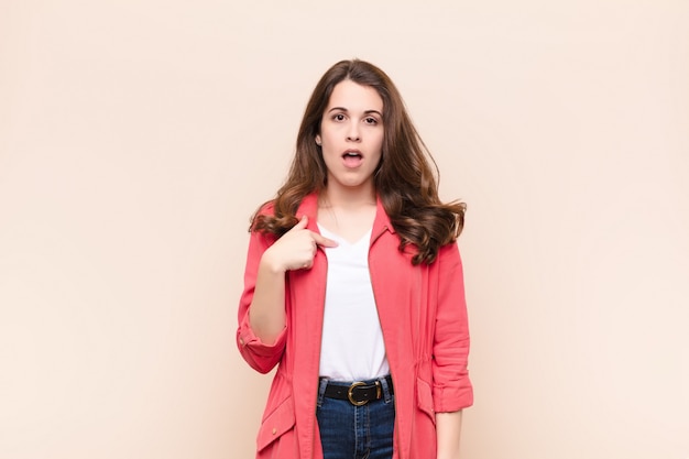 Young pretty woman looking shocked and surprised with mouth wide open, pointing to self against beige wall