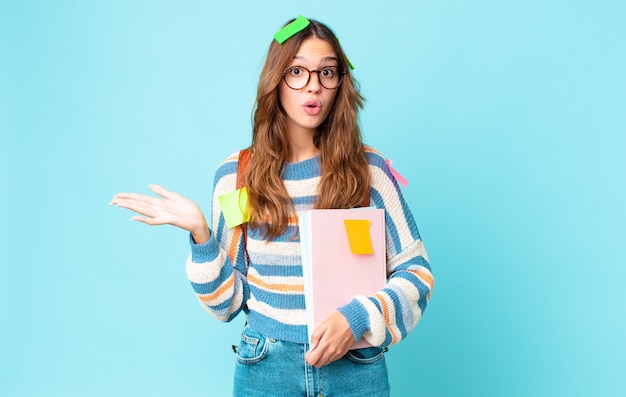 Young pretty woman looking surprised and shocked, with jaw dropped holding an object with a bag and holding books