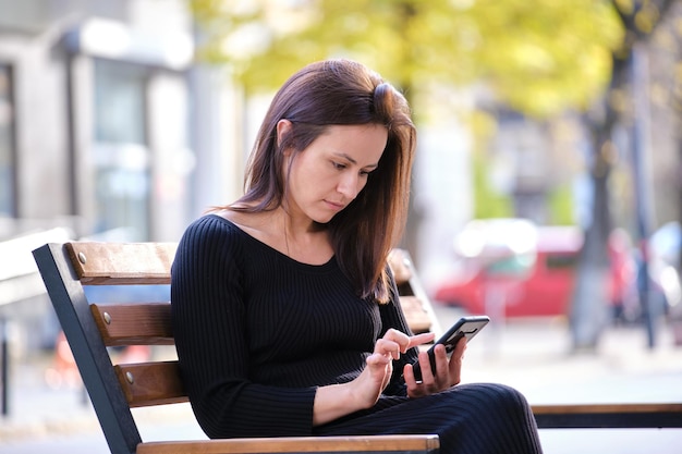 Young pretty woman messaging on mobile phone on warm summer day sitting on a city street bench outdoors