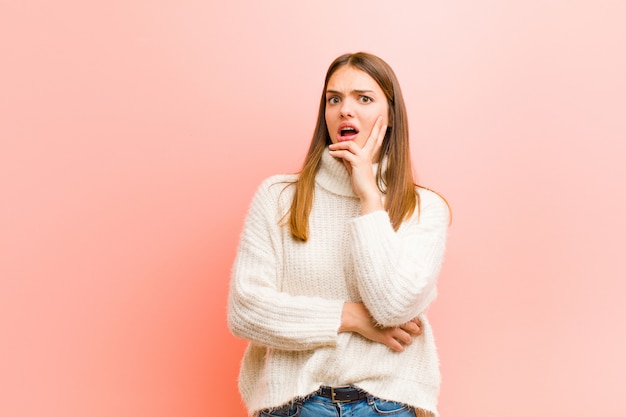 Young pretty woman open-mouthed in shock and disbelief, with hand on cheek and arm crossed, feeling stupefied and amazed against pink wall