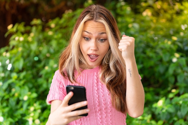 Young pretty woman at outdoors using mobile phone and doing victory gesture