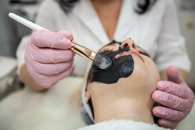 Young pretty woman receive black cosmetic facial mask at beauty salon