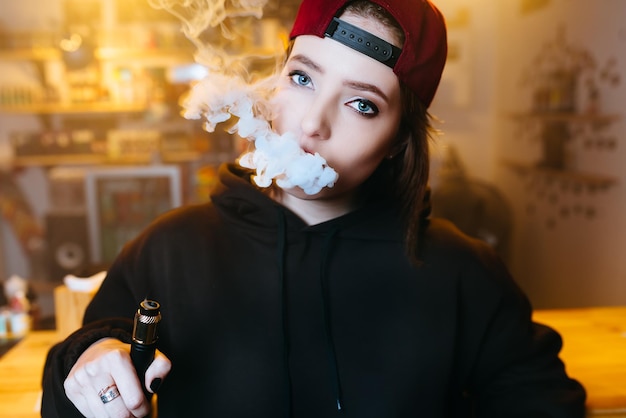 Young pretty woman in red cap smoke an electronic cigarette at the vape shop hiphop style closeup