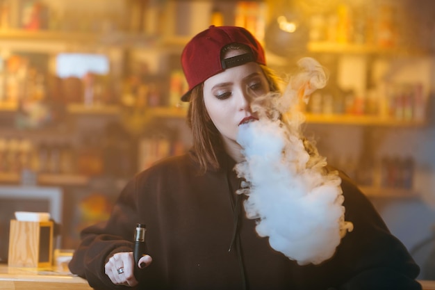 Photo young pretty woman in red cap smoke an electronic cigarette at the vape shop hiphop style closeup