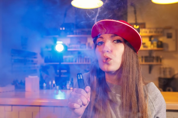 Young pretty woman in red cap smoke an electronic cigarette at the vape shop