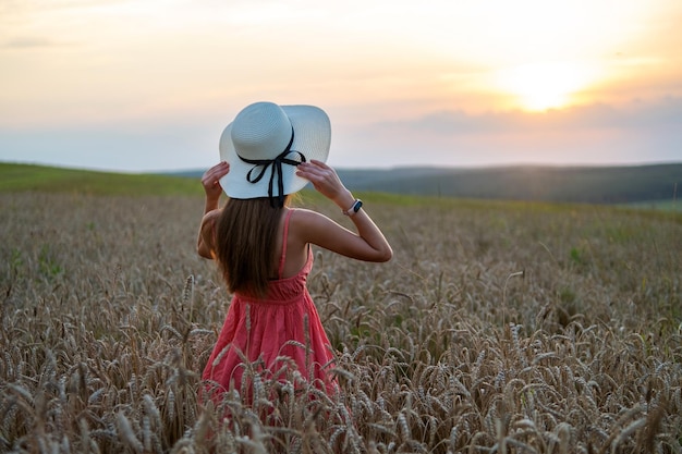 Photo young pretty woman in red summer dress and straw hat standing on yellow farm field with ripe golden wheat enjoying warm evening.