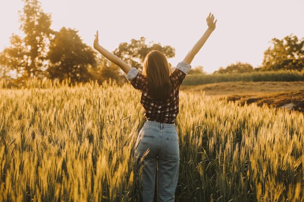 Young pretty woman in red summer dress and straw hat walking on yellow farm field with ripe golden wheat enjoying warm evening