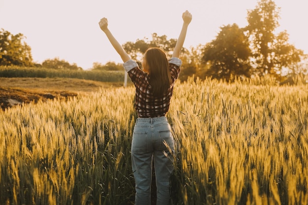 Young pretty woman in red summer dress and straw hat walking on yellow farm field with ripe golden wheat enjoying warm evening