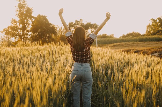 Young pretty woman in red summer dress and straw hat walking on yellow farm field with ripe golden wheat enjoying warm evening