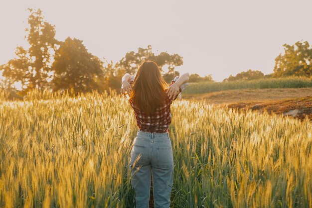 Young pretty woman in red summer dress and straw hat walking on yellow farm field with ripe golden wheat enjoying warm evening