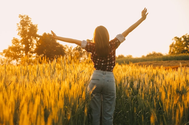 Young pretty woman in red summer dress and straw hat walking on yellow farm field with ripe golden wheat enjoying warm evening