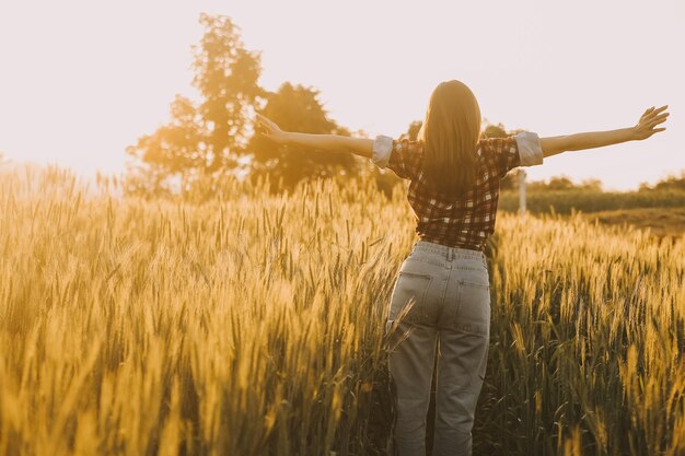 Young pretty woman in red summer dress and straw hat walking on yellow farm field with ripe golden wheat enjoying warm evening