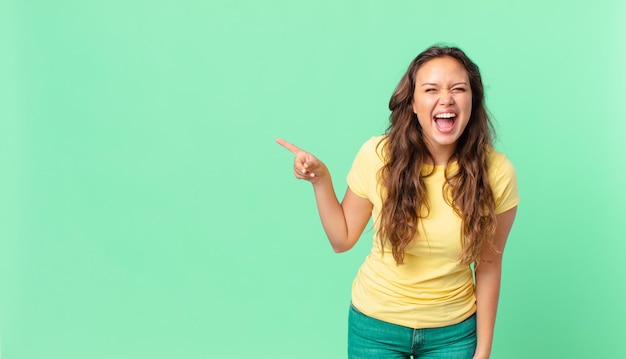 Young pretty woman shouting aggressively, looking very angry and pointing to the copy space