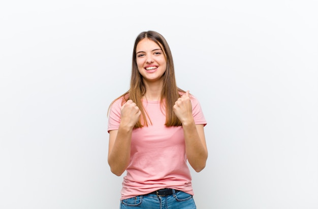 Young pretty woman shouting triumphantly, laughing and feeling happy and excited while celebrating success against white wall