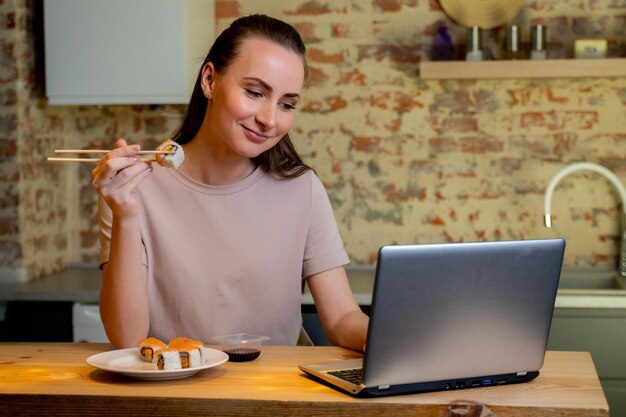 Young pretty woman sitting at her kitchen eat sushi at table home working with a laptop