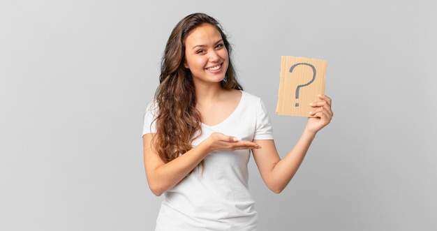 Young pretty woman smiling cheerfully, feeling happy and showing a concept and holding a question mark sign