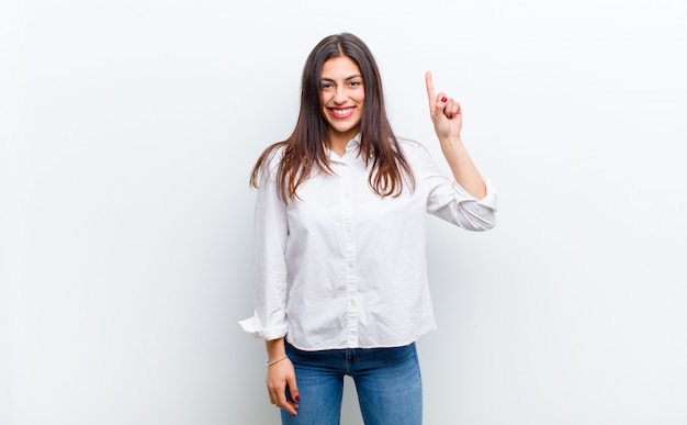 Young pretty woman smiling cheerfully and happily, pointing upwards with one hand to copy space isolated against white wall