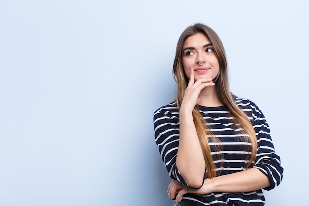 Photo young pretty woman smiling happily and daydreaming or doubting, looking to the side against soft blue wall