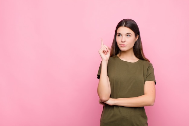 Young pretty woman smiling happily and looking sideways, wondering, thinking or having an idea over pink wall