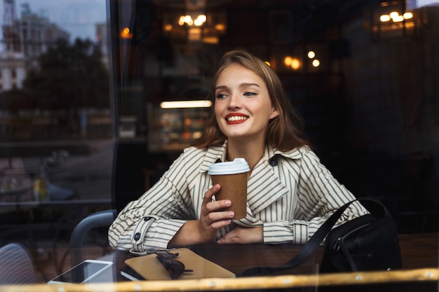 Young pretty woman in striped trench coat holding cup to go in hand joyfully 