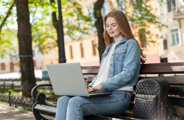 Young pretty woman student or freelancer using laptop while sitting on a bench in the city