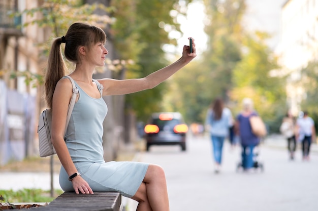 Young pretty woman taking selfie with mobile phone on warm summer day sitting on a city street bench outdoors.