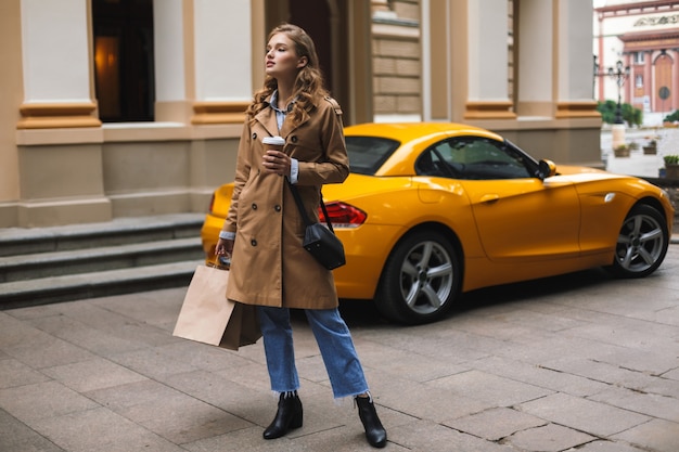 Young pretty woman in trench coat with little black cross bag holding cup of coffee to go and shopping bags in hands while thoughtfully 