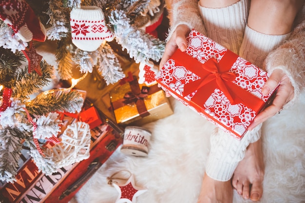 Young pretty woman in warm cozy clothes is sitting on the light wooden floor in her bright house near the christmas tree and holds gift box in her hands.