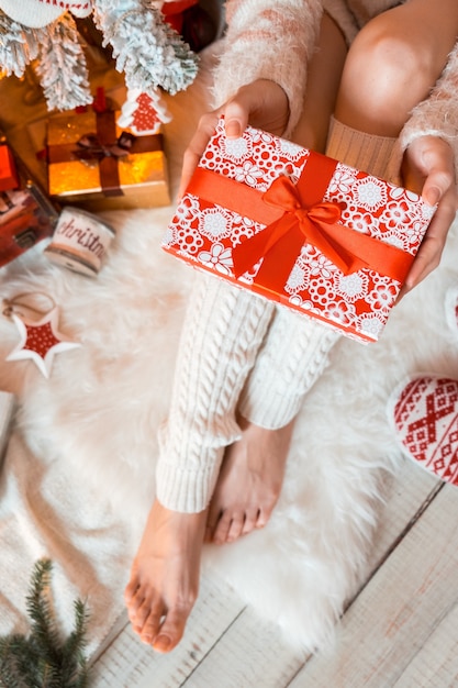 Young pretty woman in warm cozy clothes is sitting on the light wooden floor in her bright house near the christmas tree and holds gift box in her hands.