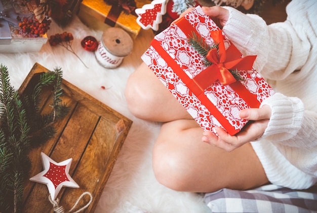 Young pretty woman in warm cozy clothes is sitting on the light wooden floor in her bright house near the christmas tree and holds gift box in her hands.