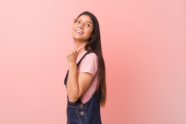 Young pretty woman wearing a jeans dungaree points with thumb finger away, laughing and carefree.