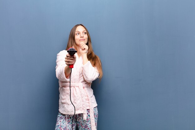 Young pretty woman wearing pajamas and holding a hairdresser against blue wall with a copyspace