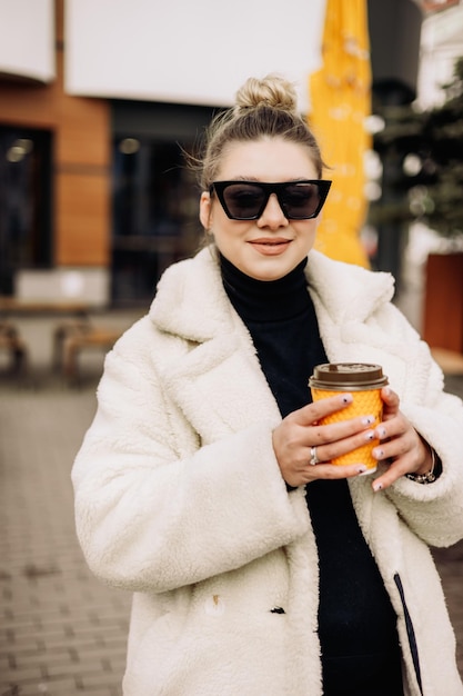 A young pretty woman in a white fur coat and sunglasses with coffee walks around the city Stylish young girl