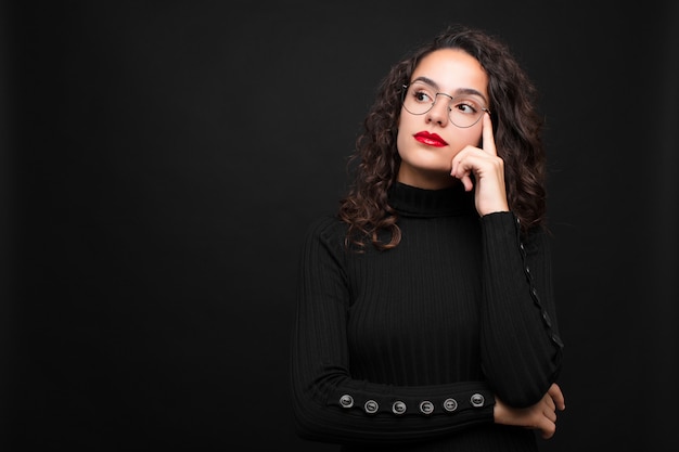 Young pretty woman with a concentrated look, wondering with a doubtful expression, looking up and to the side against black wall.