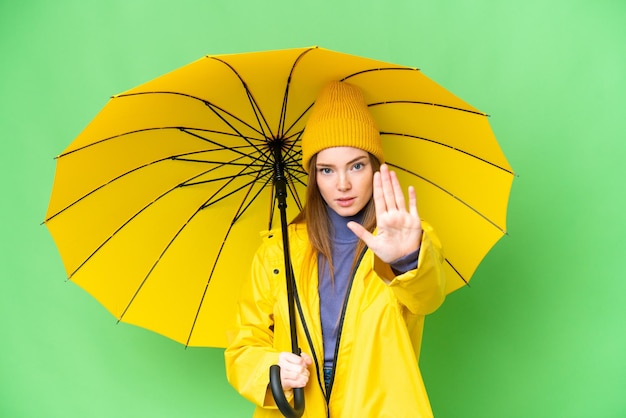 Young pretty woman with rainproof coat and umbrella over isolated chroma key background making stop gesture