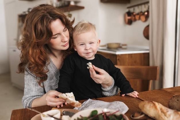 Young pretty woman with red hair in knitted sweater feeding her little smiling son that joyfully 