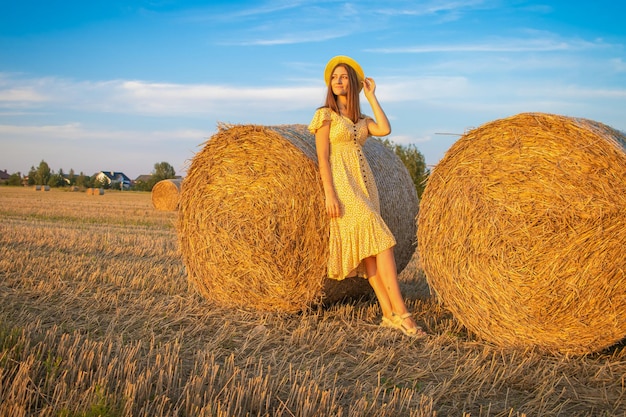 Young pretty woman in yellow dress and hat close up on the field background with haystacks at sunset time