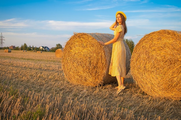 Young pretty woman in yellow dress and hat close up on the field background with haystacks at sunset time