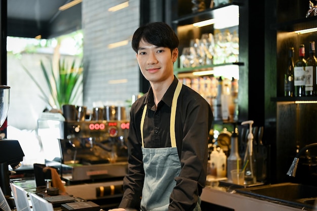 Young professional Asian male barista standing in the cafe and smiling to the camera