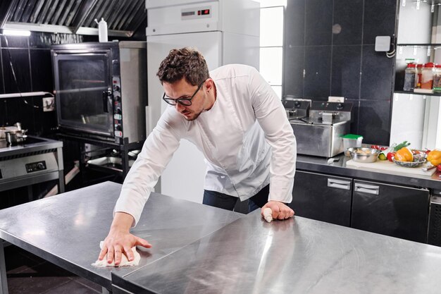 Young professional cook in uniform wiping kitchen counter in kitchen after cooking