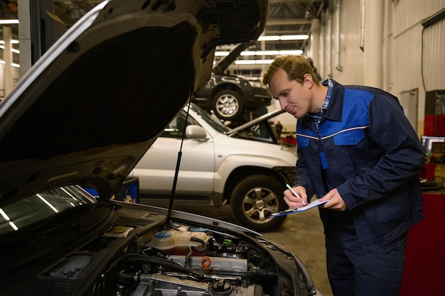 Photo young professional mechanic writes on clipboard while inspecting the engine of an automobile wit open hood in auto service repair shop